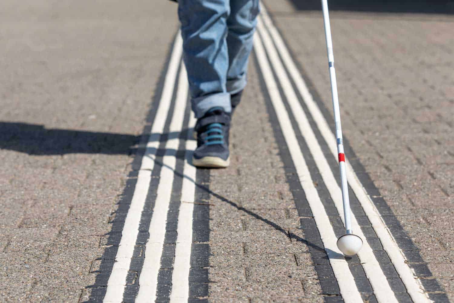 Beine einer sehbeeinträchtigten Person mit Blindenstock neben weissen Bodenleitlinien für Blinde.
