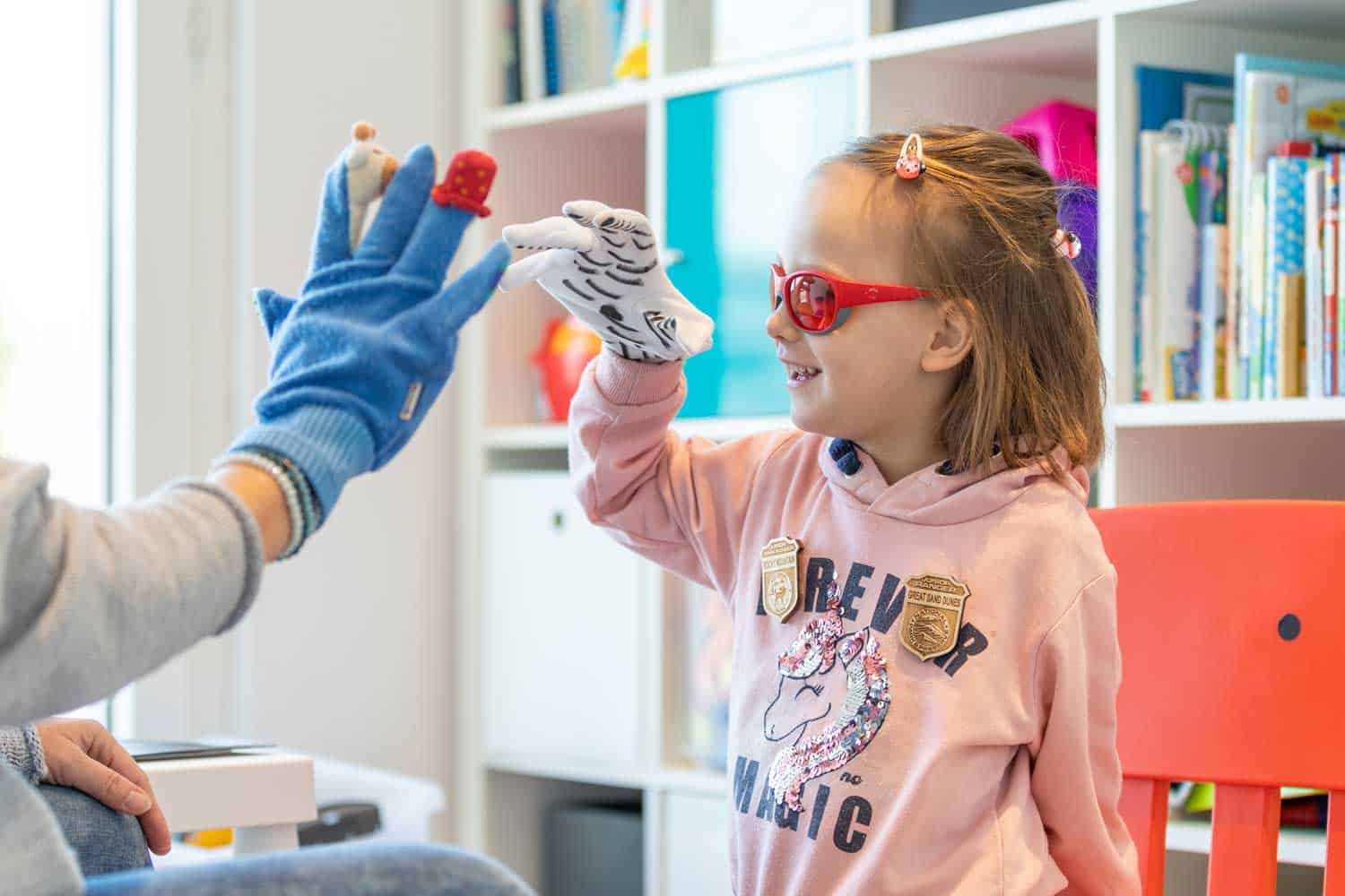 Junges Mädchen mit Seh- und Blindheit und Sehbeeinträchtigung mit Mehrfachbeeinträchtigungen lacht beim Spielen mit Fingerpuppen zusammen mit einer Therapeutin.