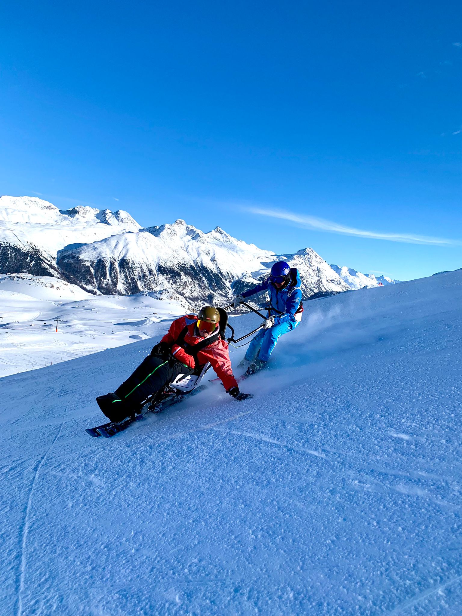 Schülerin mit Monoski und Skilehrperson auf der Piste. Im Hintergrund ist ein beeindruckendes Bergpanorama zu sehen.